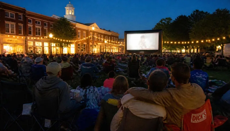 Members watching an outdoor movie night on a town green at dusk, wrapped in blankets